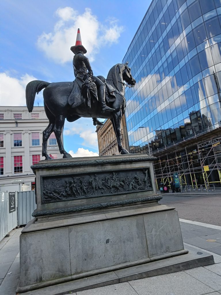 A statue of a man on horseback wearing a traffic cone on his head stands proudly with modern buildings in the background—an iconic sight near some of the must visit museums in Glasgow.
