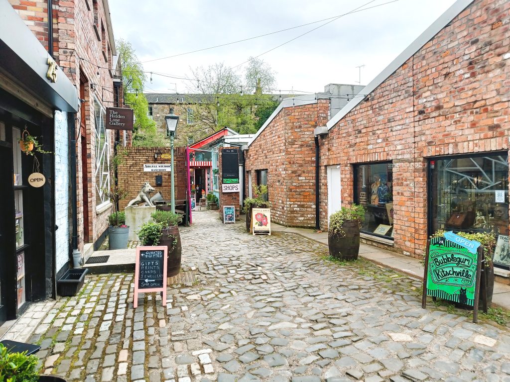 A cobblestone alley lined with brick shops, chalkboard signs, and potted plants on a cloudy day—just one of the 10x Best things to do in Glasgow.