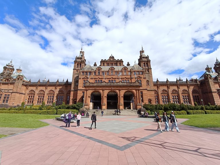 People walk in front of the Kelvingrove Art Gallery, a stunning red-brick museum in Glasgow, with its towers rising under a partly cloudy blue sky.