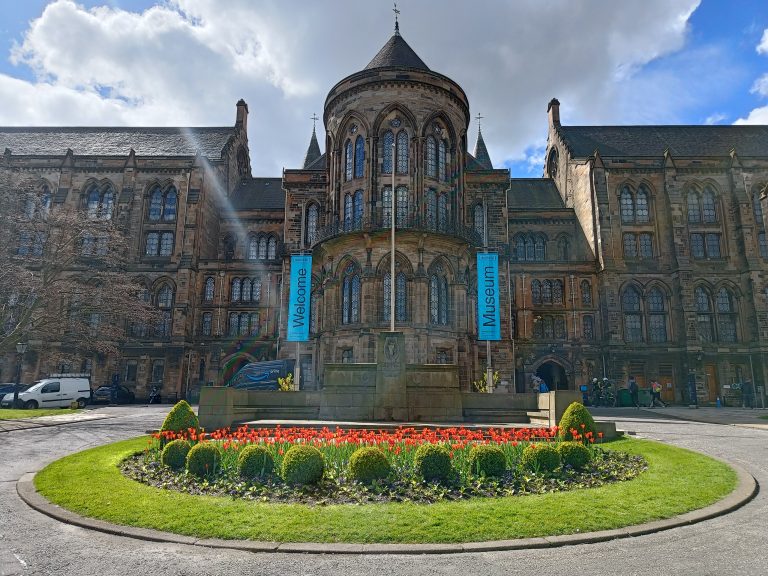 Historic stone building with “Welcome” and “Museum” banners, round flowerbed and red tulips in front—one of the must visit museums among Glasgow museums.
