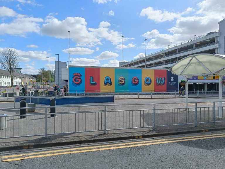 A colorful "GLASGOW" sign on a wall outside a parking garage under a partly cloudy sky highlights the vibrant spirit of travel in Glasgow.