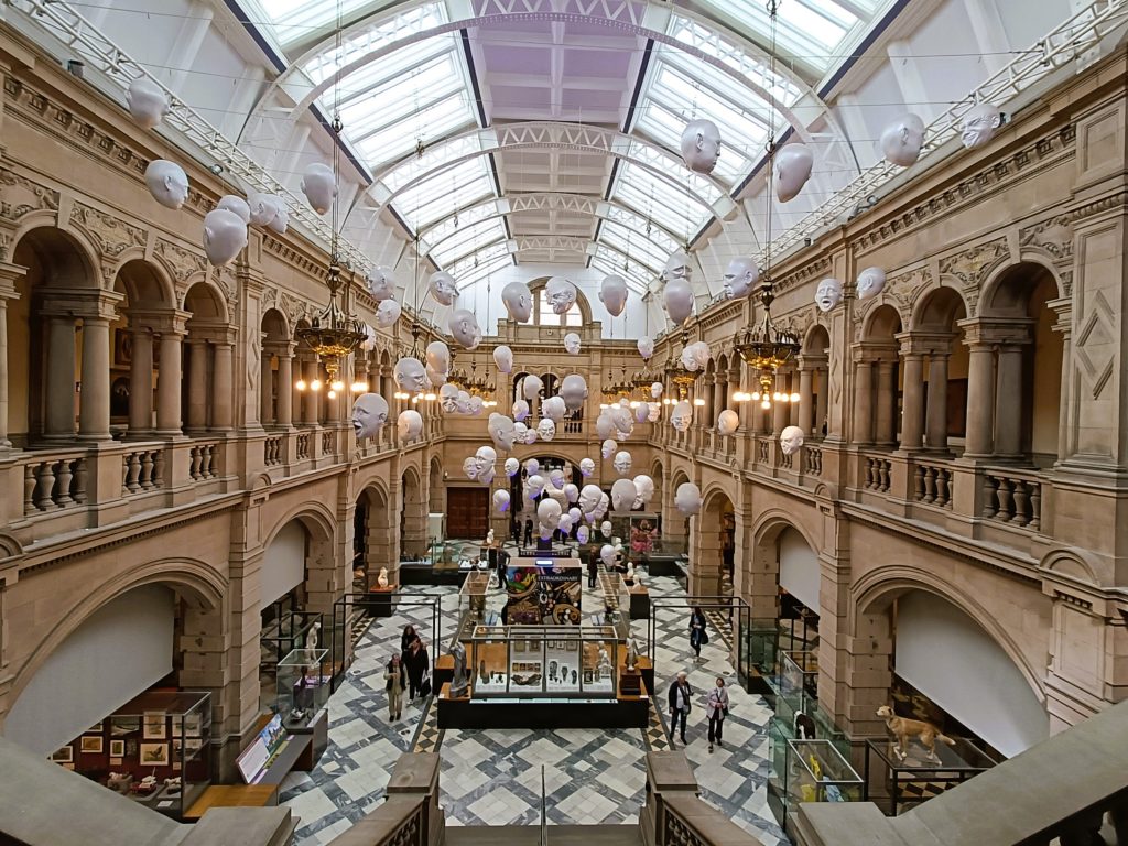 A spacious hall at the Kelvingrove Art Gallery in Glasgow, featuring arched balconies and white balloon faces suspended from the ceiling.
