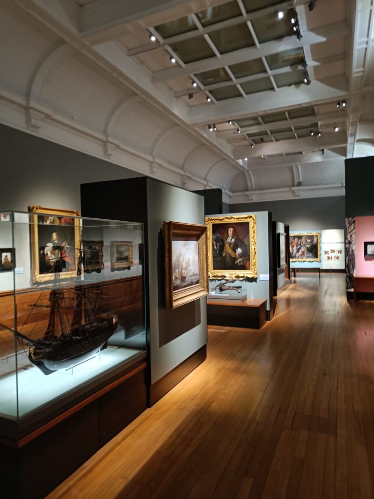 A museum gallery in Glasgow with paintings on the walls and model ships in glass cases on a polished wooden floor, reminiscent of the Kelvingrove Art Gallery’s elegant displays.
