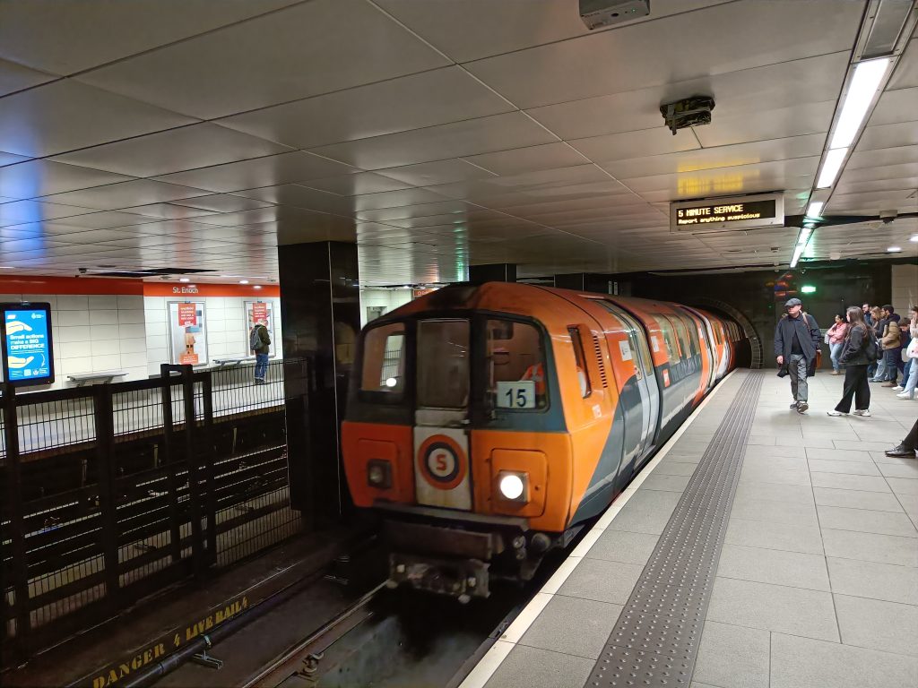 An orange subway train arrives at an underground platform with people waiting nearby, showcasing a popular way of getting around Glasgow.