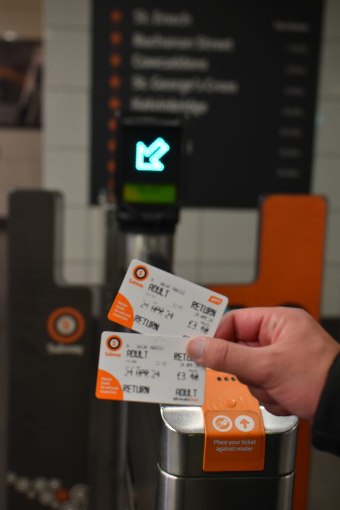 Hand holding two train tickets near a turnstile with a green arrow and a place for ticket scanning—an easy example of how to get around in Glasgow.