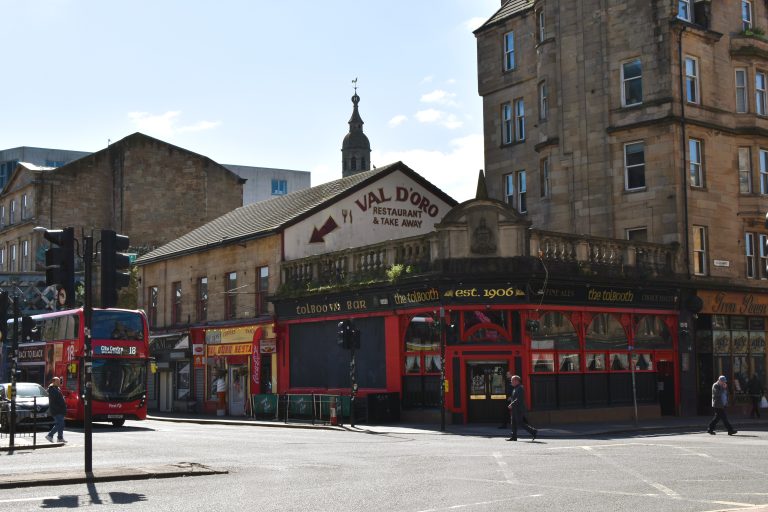 A red double-decker bus passes Val D’Oro restaurant and tall stone buildings on a city street—an iconic scene among the 10x Best things to do in Glasgow.