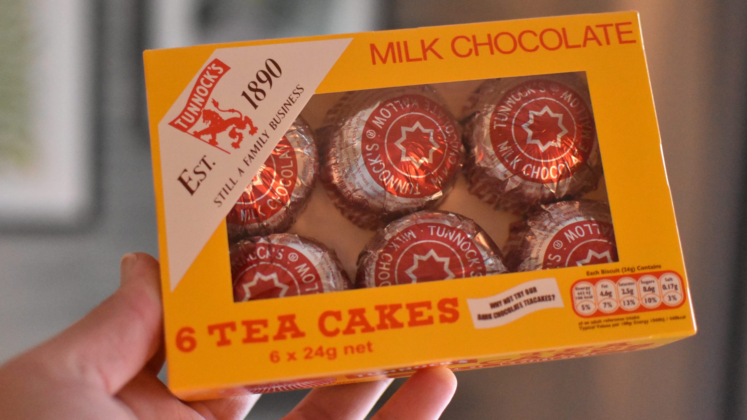 A hand holds a box of Tunnock's Milk Chocolate Tea Cakes, a classic food souvenir from Glasgow, containing six individually wrapped cakes.