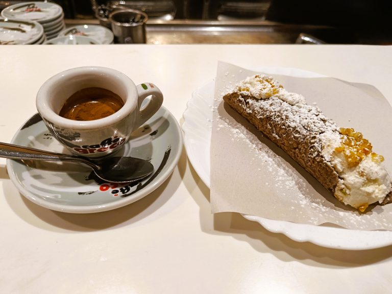 A cup of coffee and a cannoli with powdered sugar on a white plate sit on the counter—an inviting taste of Naples perfect for breakfast.