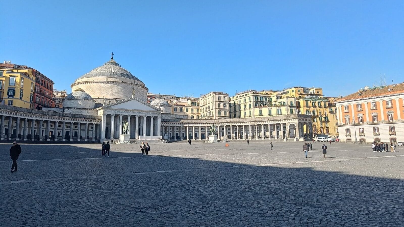 Large open square with few people, a domed church, columns, and colorful buildings under a clear blue sky—one of the top things to do in Naples and a must-see among Naples attractions.