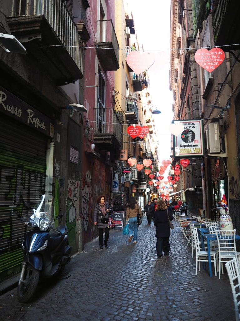 Narrow cobblestone street with heart decorations, people walking, and outdoor café seating on the right—a charming spot for Naples travel and discovering unique Naples attractions.