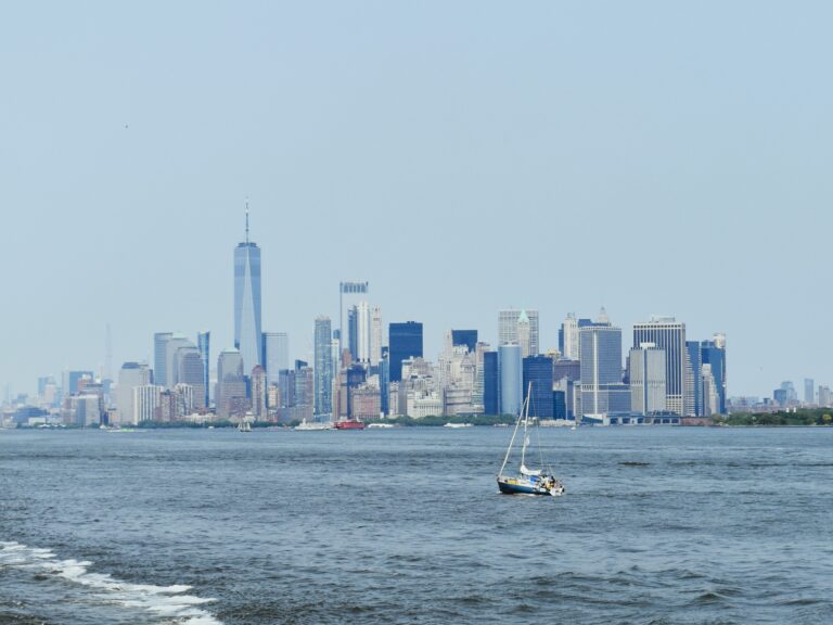 A sailboat on the water glides past the skyline, showcasing the iconic boroughs of New York City under a clear sky.