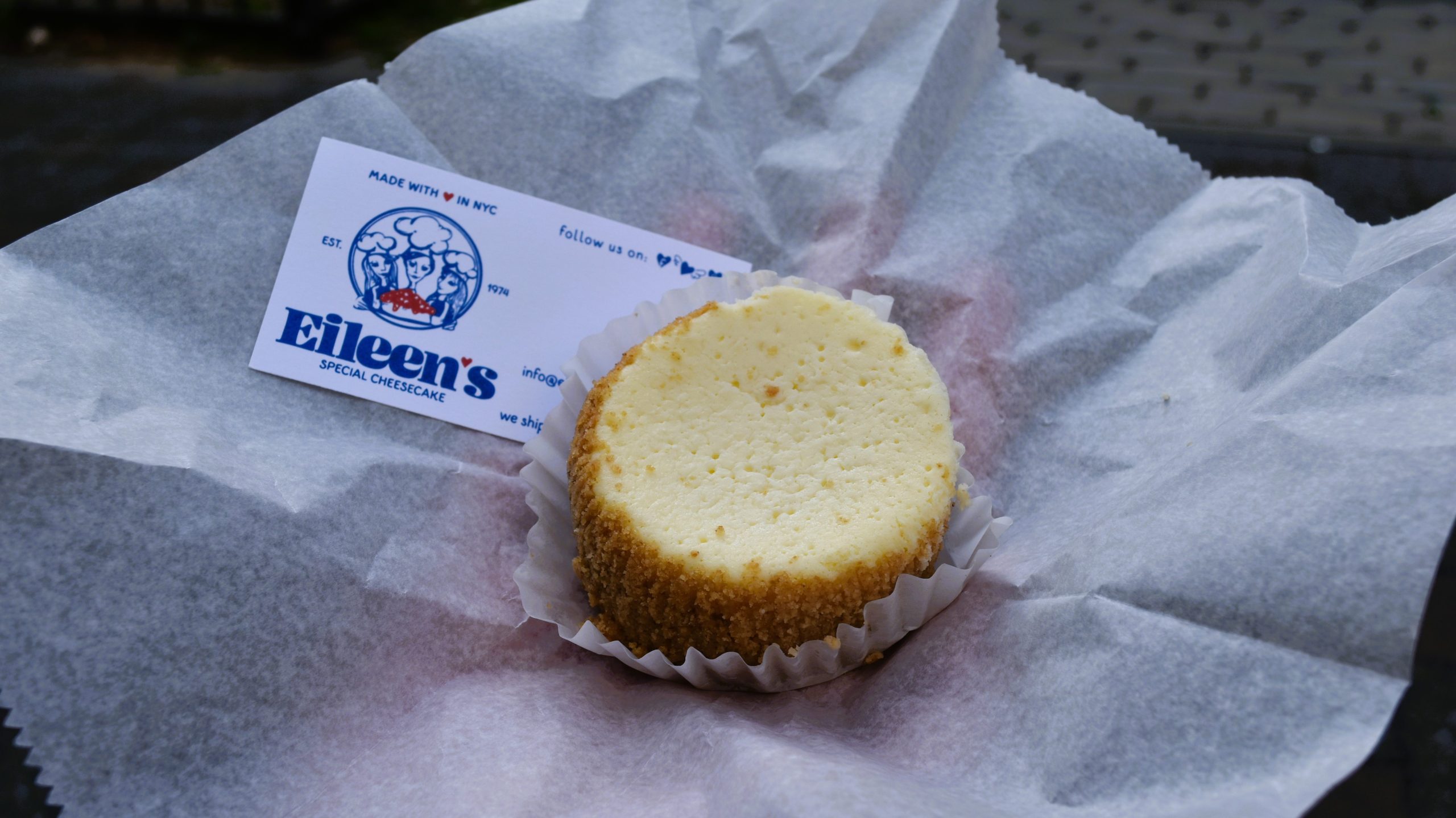 Small cheesecake on wax paper with an Eileen’s Special Cheesecake business card beside it, showcasing a classic taste of food in New York.