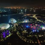 Aerial night view of Gardens by the Bay in Singapore, illuminated with colorful lights near the waterfront.