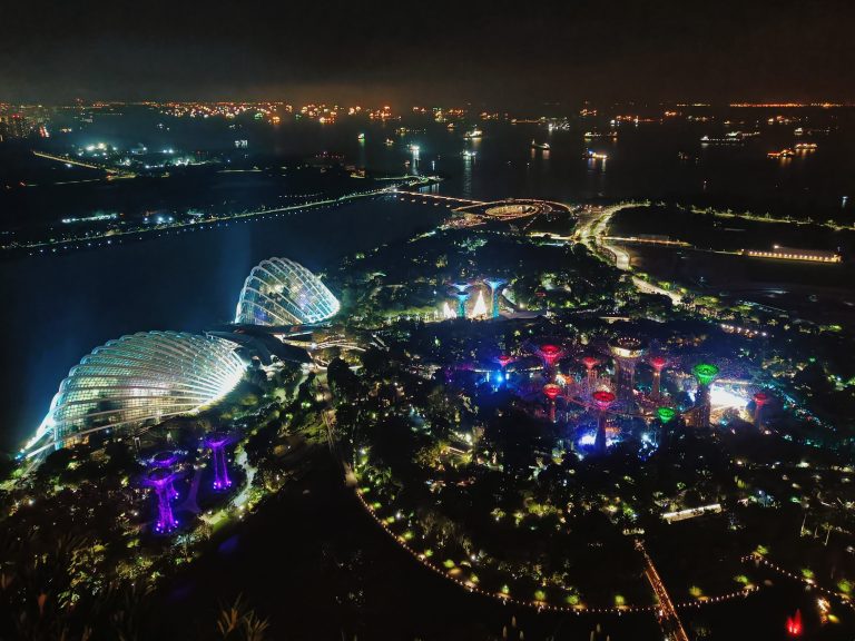 Aerial night view of Gardens by the Bay in Singapore, illuminated with colorful lights near the waterfront.