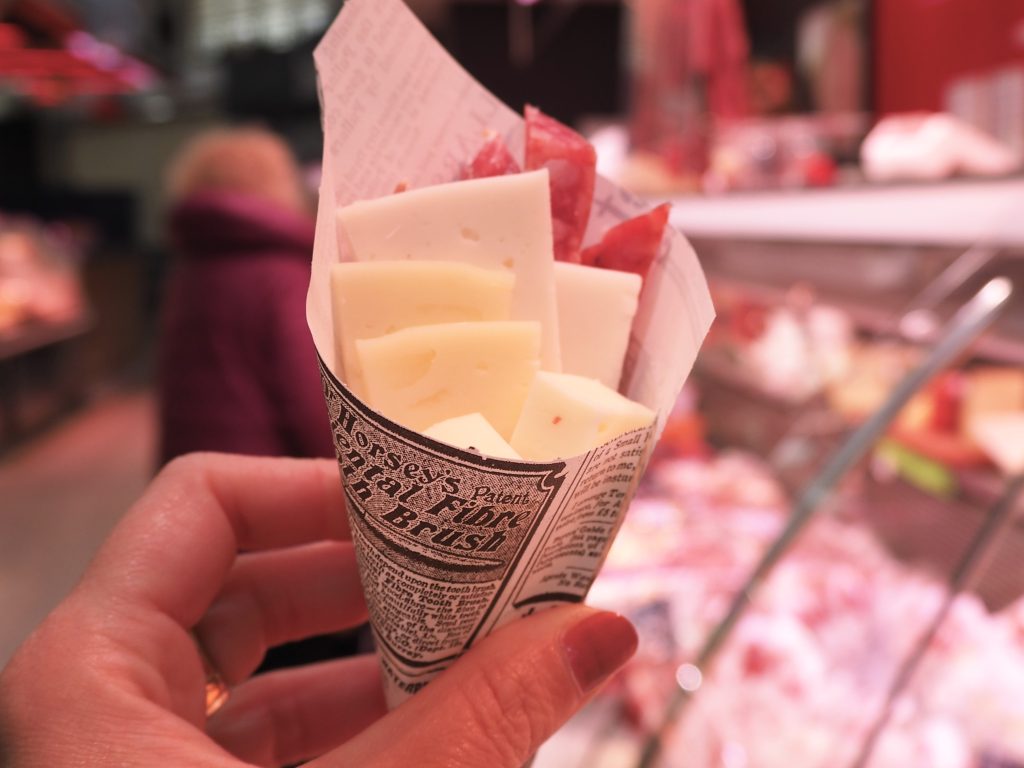 A hand holds a paper cone filled with cheese slices and cured meat in a deli setting.