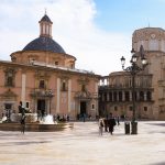 People walk around a plaza with a fountain, historic buildings, and a cathedral under a partly cloudy sky.