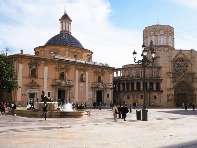People walk around a plaza with a fountain, historic buildings, and a cathedral under a partly cloudy sky.