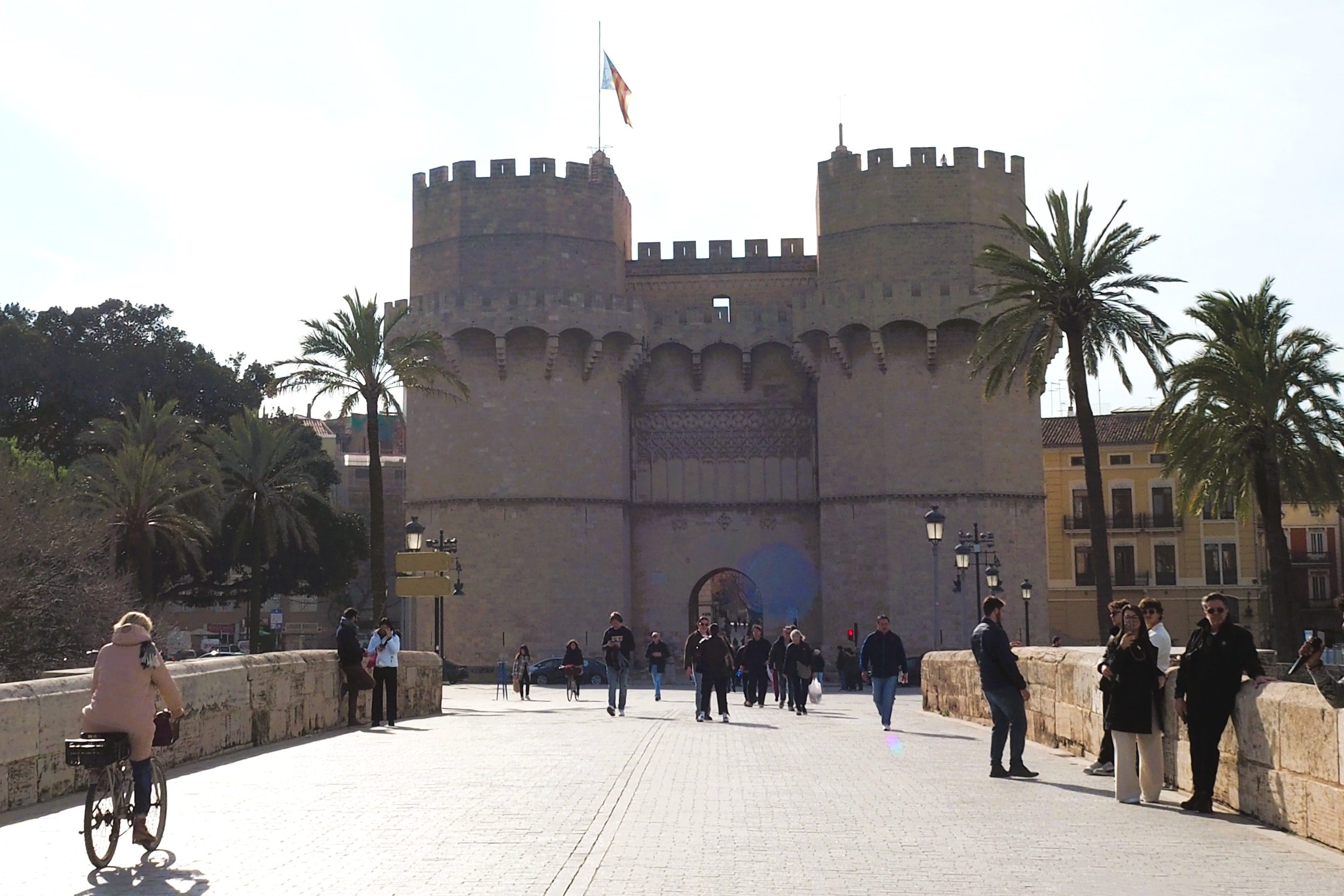 People walk and cycle toward a historic stone gate with towers, palm trees, and a flag—an iconic sight in one of the Popular Neighborhoods in Valencia.