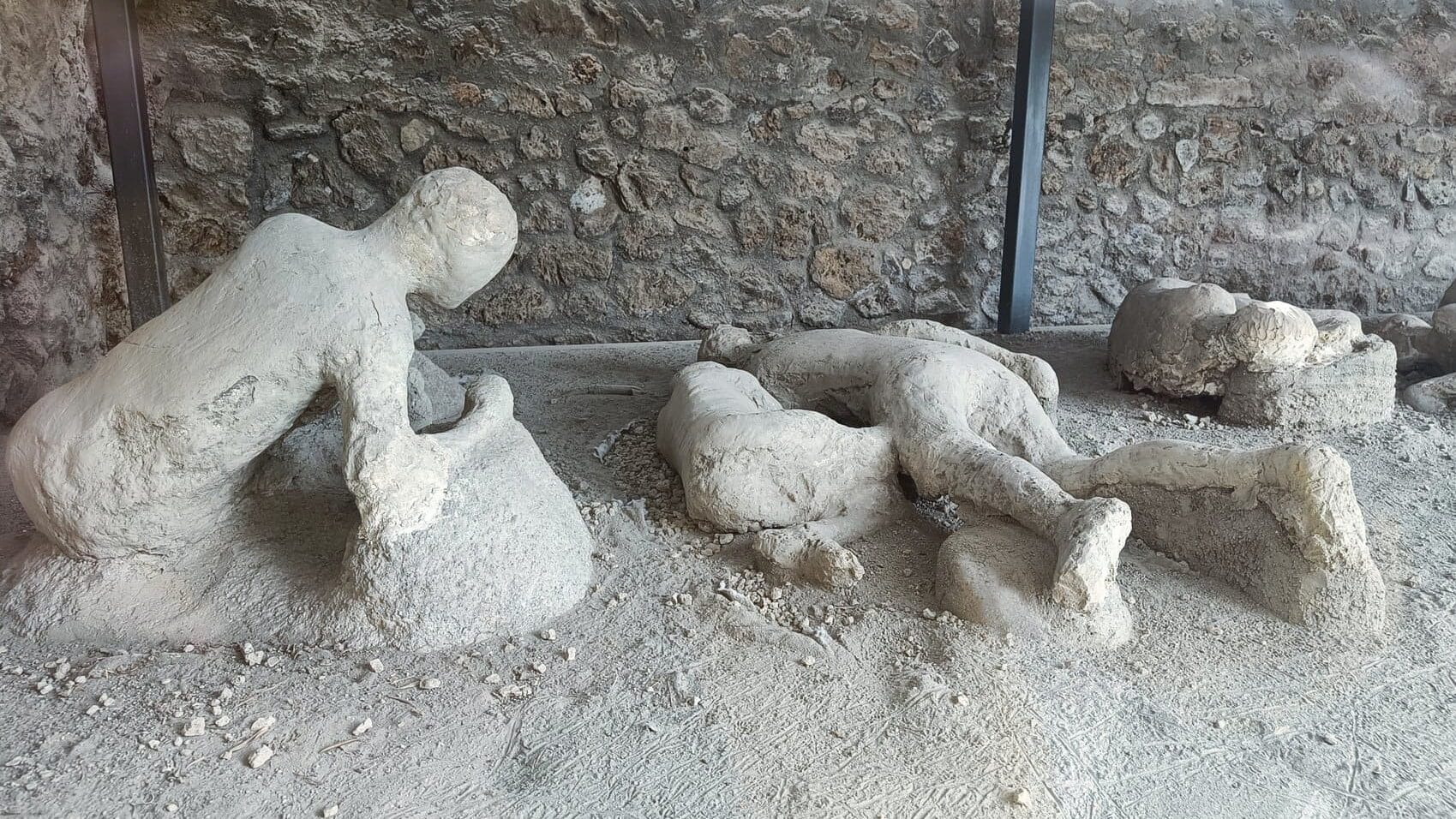 Plaster casts of human figures preserved in ash at an archaeological site with a stone wall in the background, as seen in some of 4x the best museums in Naples.