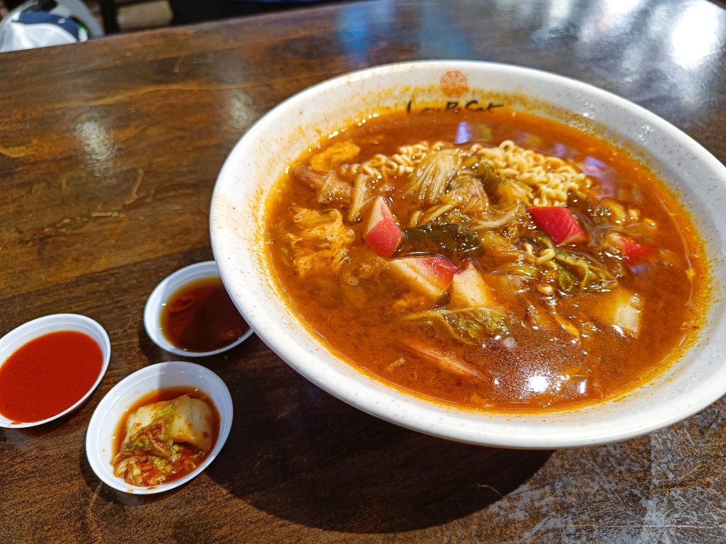 A bowl of spicy ramen soup with vegetables, served with kimchi and dipping sauces on a wooden table.