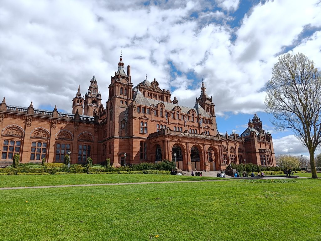 Red-brick Victorian building with towers, surrounded by green lawn and trees under a partly cloudy sky—one of the 10x Best things to do in Glasgow for architecture lovers.