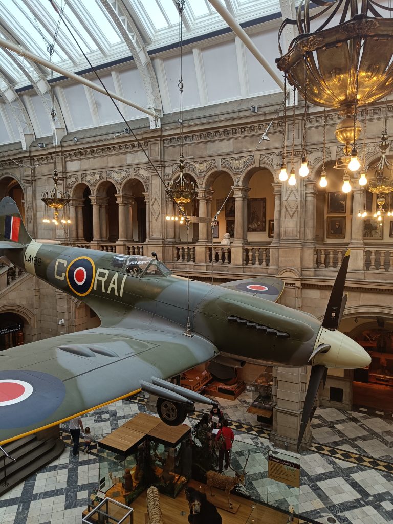 A vintage fighter plane is displayed indoors at the Kelvingrove Art Gallery, a grand museum in Glasgow with arched balconies and sparkling chandeliers.