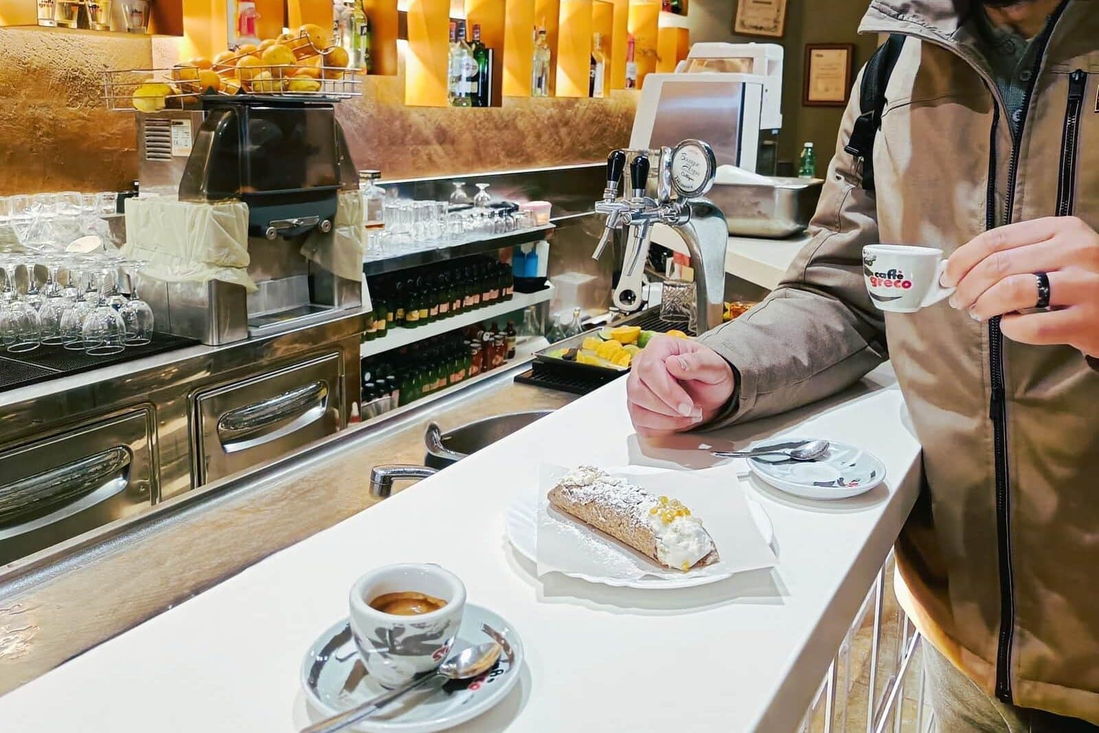 Person at a café counter in Naples with a fresh espresso, a breakfast pastry on a plate, and shelves of drinks in the background. Breakfast and Coffee in Naples