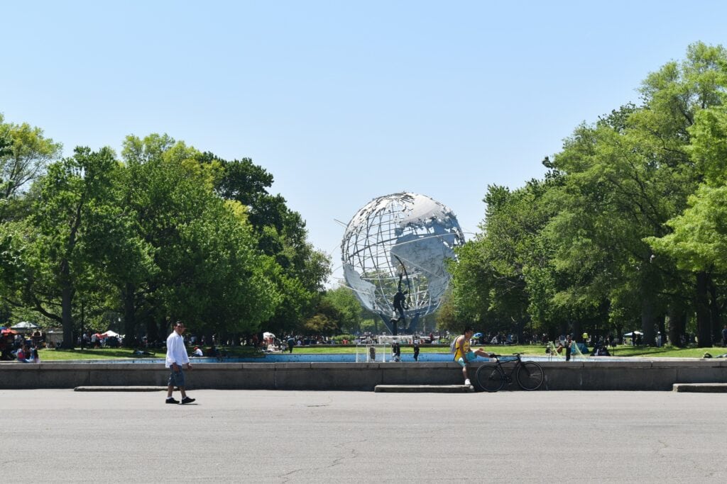 People walk and relax near the Unisphere globe sculpture, surrounded by trees in a city park—a landmark symbolising unity within the boroughs of New York City—on a sunny day.
