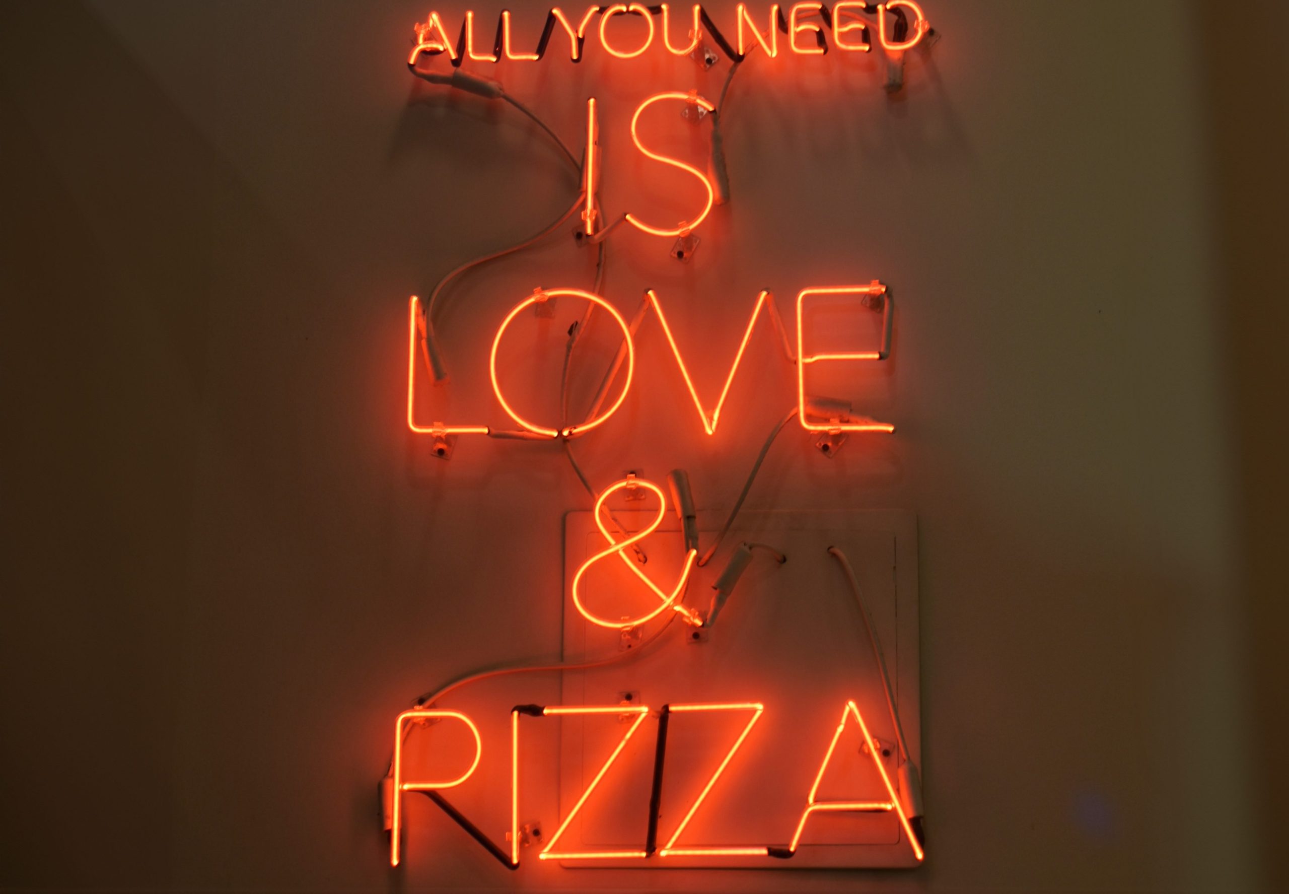 Neon sign reads: "ALL YOU NEED IS LOVE & PIZZA" in orange-red letters on a wall, capturing the spirit of food in New York.