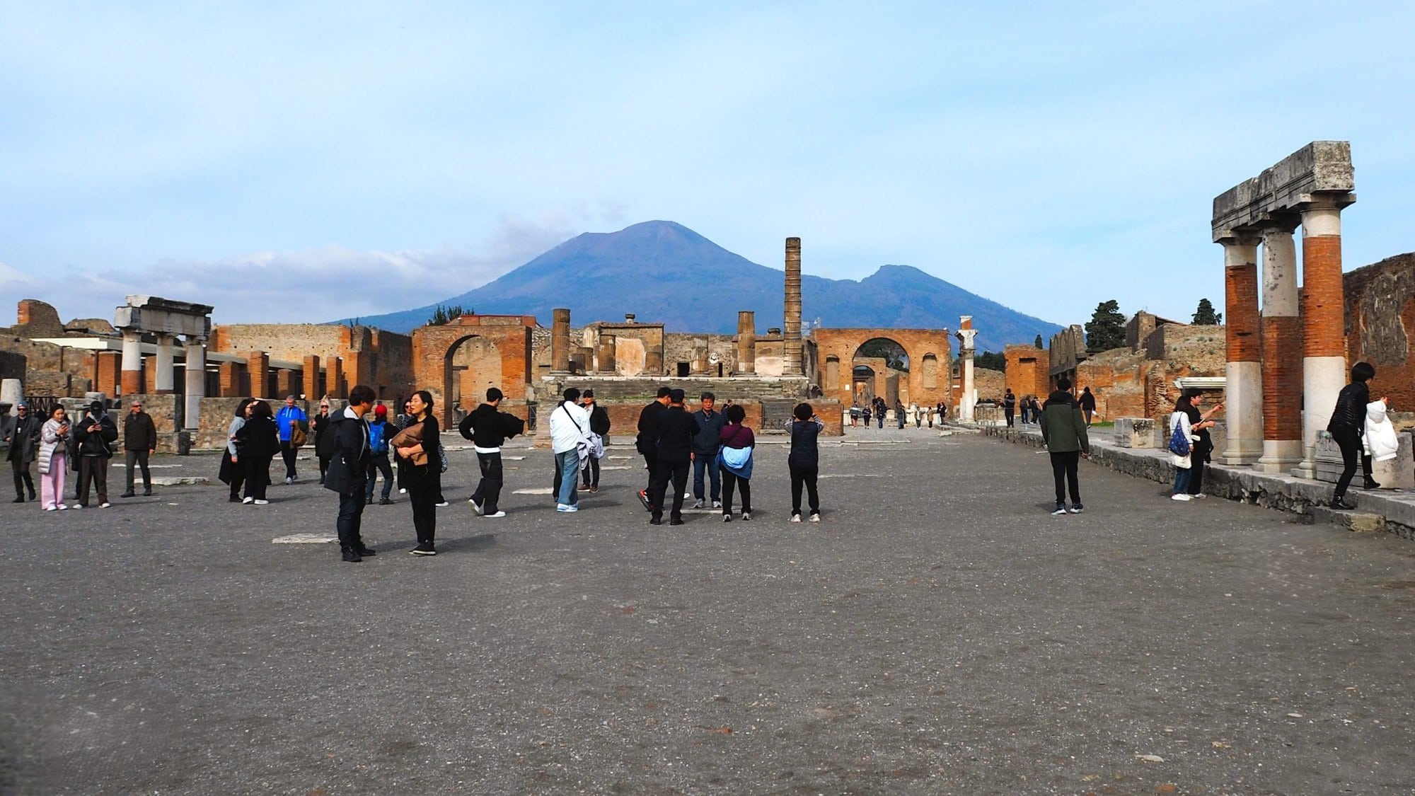 Tourists explore ancient ruins in Pompeii with Mount Vesuvius visible in the background under a blue sky, making it one of the best day trips from Naples.
