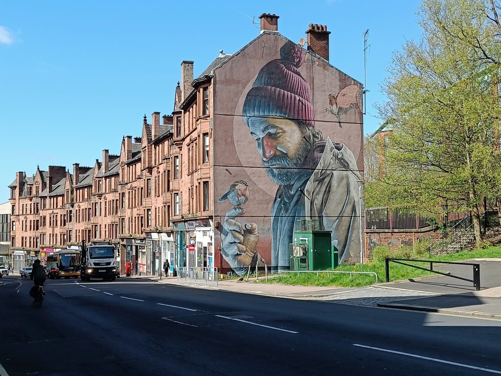 A mural of a man with birds is painted on the side of a building along a city street, making it one of the 10x Best things to do in Glasgow for art lovers and explorers.
