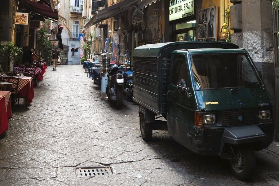 Narrow cobblestone street in one of the popular neighborhoods of Naples, with parked scooters and small vehicles beside outdoor cafes and old buildings, showcasing the lively charm typical of 6x Popular neighborhoods in Naples