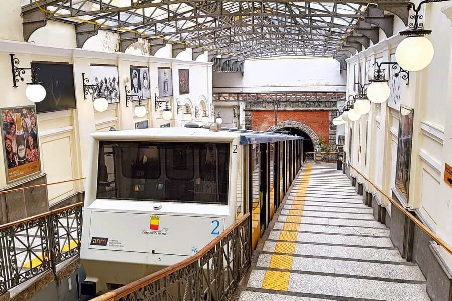A white funicular train at a covered station, with stairs and vintage lamps on either side—a charming way to explore the popular neighborhoods of Naples.