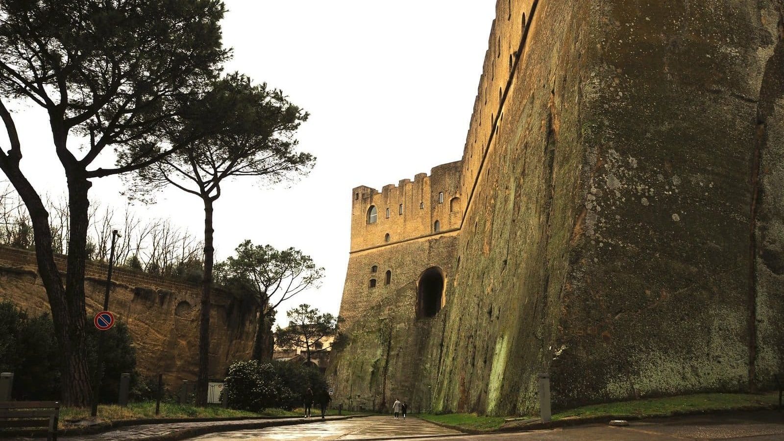 Tall ancient stone fortress walls and towers, with trees beside a wet, empty street on a cloudy day—one of the best attractions Naples has to offer for those seeking history among its atmospheric sights.