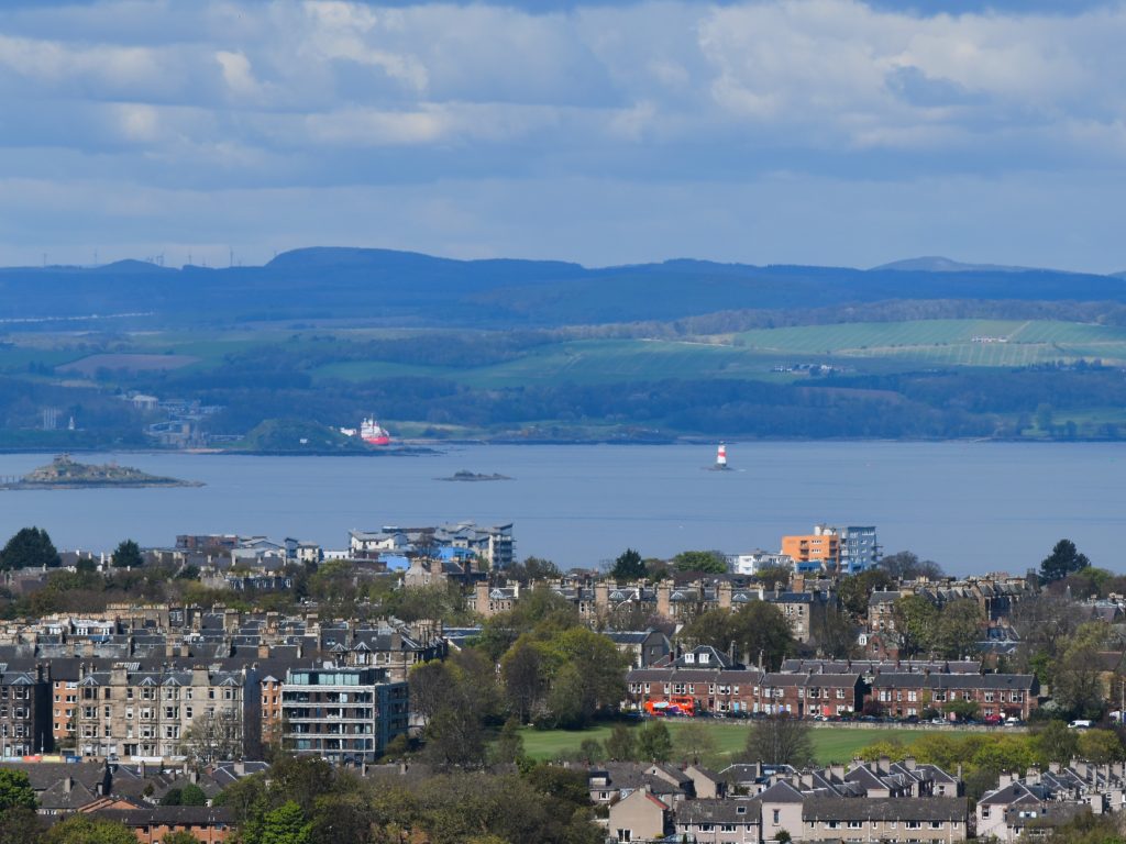 A panoramic view of a coastal town with houses, trees, water, and distant hills under a partly cloudy sky—evoking the charm that inspires searches for where to stay in Edinburgh.