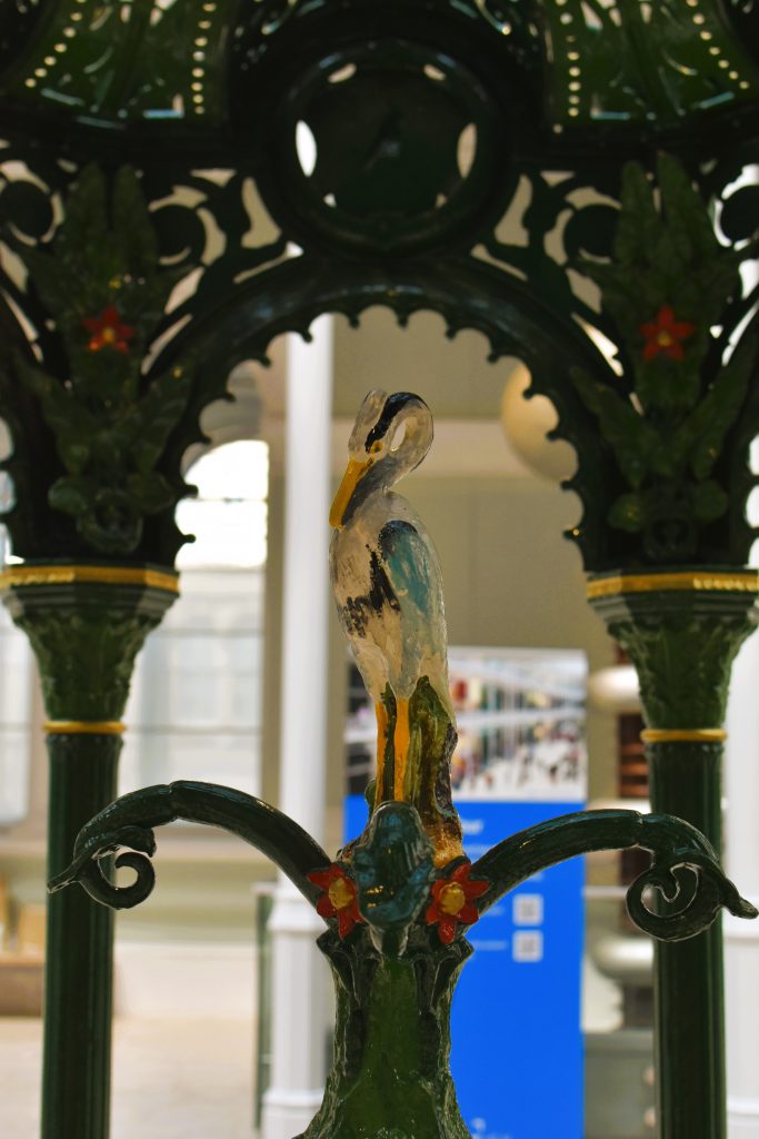 A painted bird statue sits on a green ornate stand with floral details in an indoor setting, reminiscent of unique exhibits found in the best museums Edinburgh has to offer.