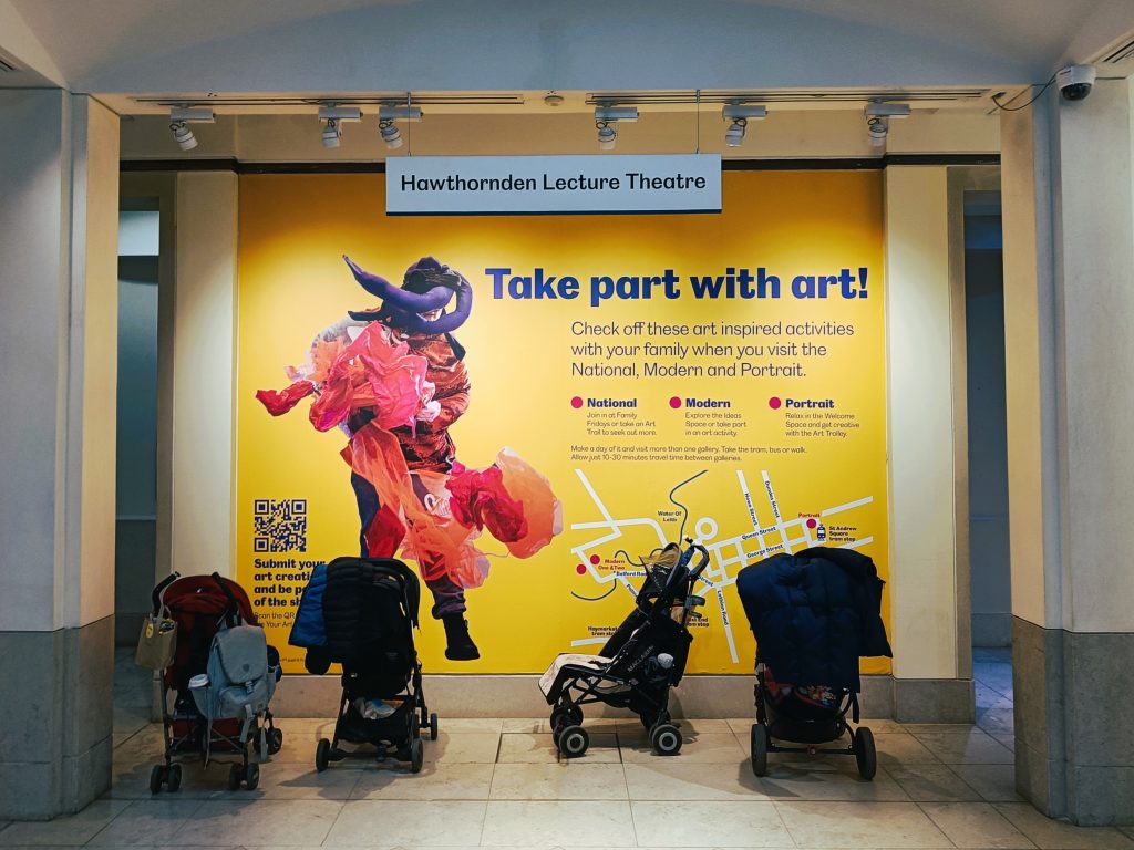 Four strollers are parked in front of a bright yellow art activity sign at Hawthornden Lecture Theatre, one of the highlights at one of the best museums Edinburgh has to offer.