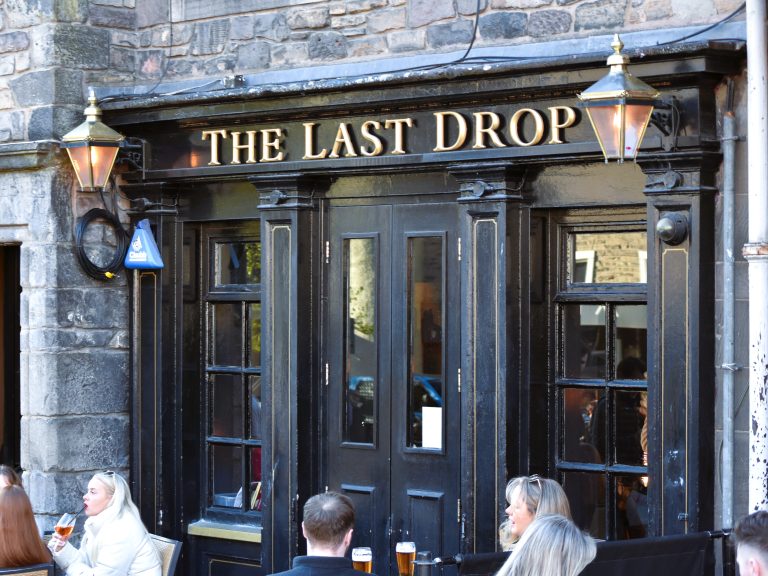 People sitting outside a pub called "The Last Drop," with drinks on tables and a stone building facade—one of the best pubs in Edinburgh.