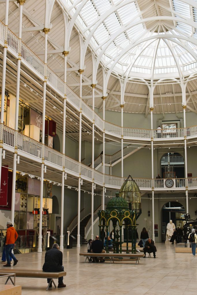 Interior of a spacious, bright museum hall with a vaulted glass ceiling, people seated and walking around exhibits—a glimpse inside one of the best museums Edinburgh has to offer.