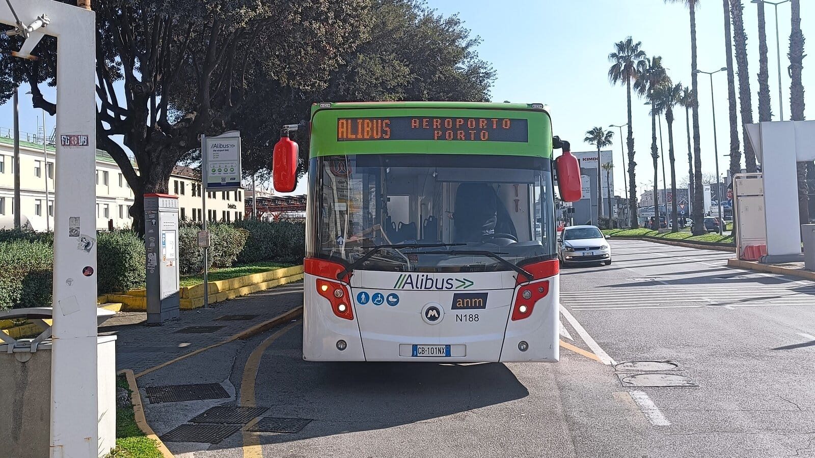 A green and white airport shuttle bus labeled "Alibus," part of Naples transportation, is parked at a sunny street stop.