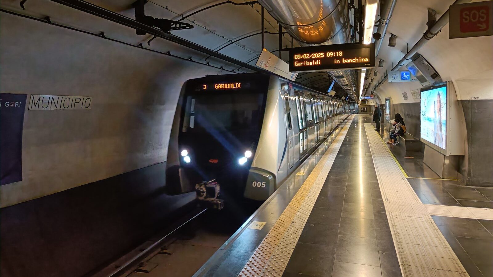 A modern subway train arrives at an underground station, offering a glimpse into efficient Naples transportation as a few people wait on the platform.
