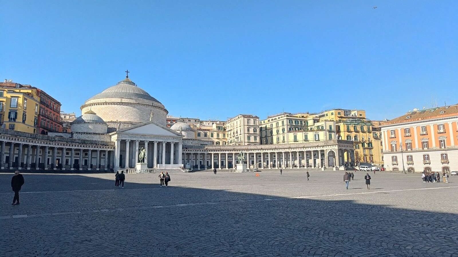 Wide plaza with people walking, a domed church, columns, and colorful buildings under a clear blue sky—this vibrant scene captures the essence of one of the popular neighborhoods in Naples.