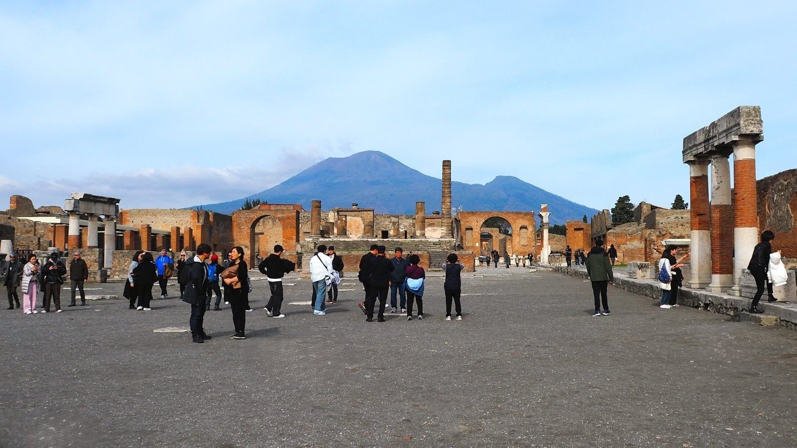 Tourists explore ancient ruins in Pompeii with Mount Vesuvius visible in the background under a blue sky, making it one of the best day trips from Naples.
