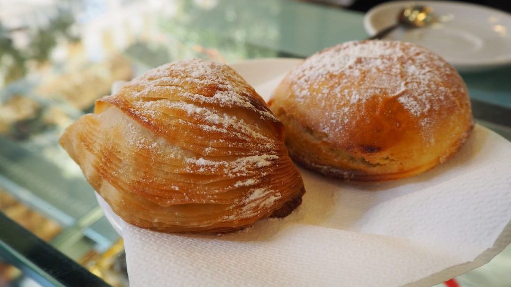 Two Italian pastries, one flaky and one round, dusted with powdered sugar on a napkin in a café.