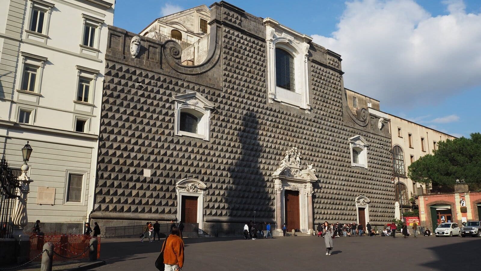 People walk in front of a large, dark stone building with a patterned facade and arched windows under a blue sky, one of the best places in Naples to admire historic architecture.