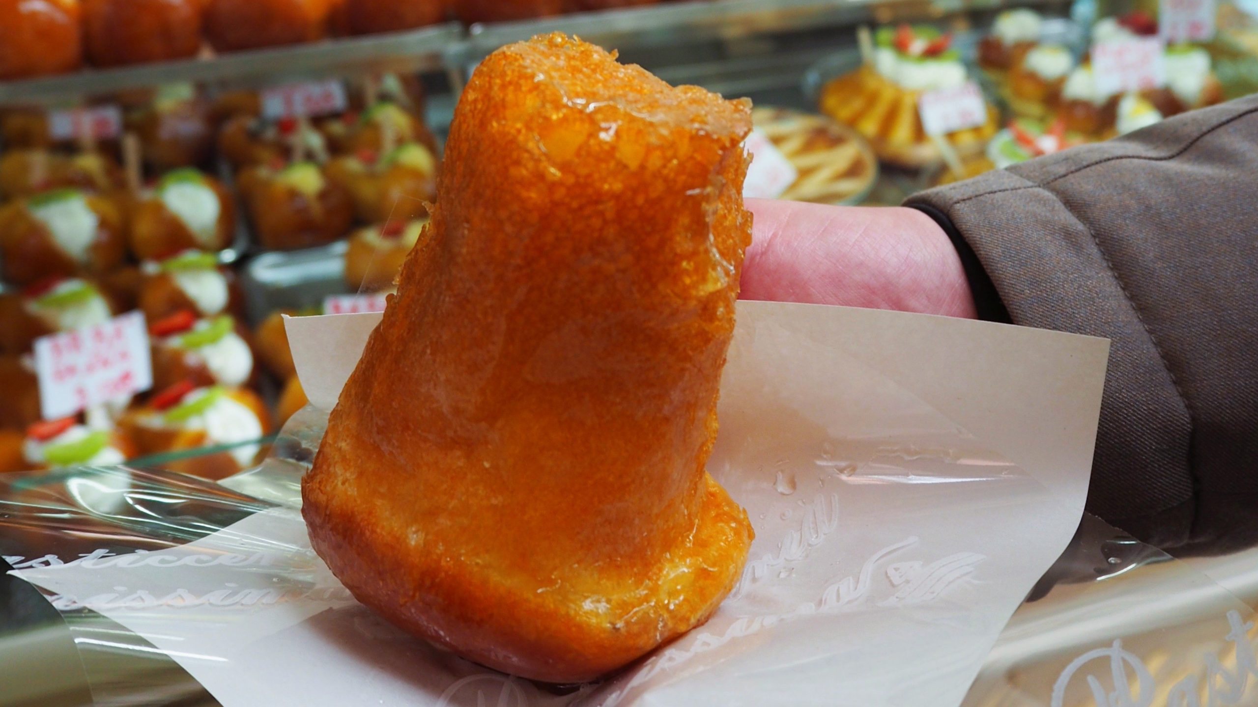 A hand holding a shiny, golden pastry wrapped in paper at a bakery display with assorted desserts.