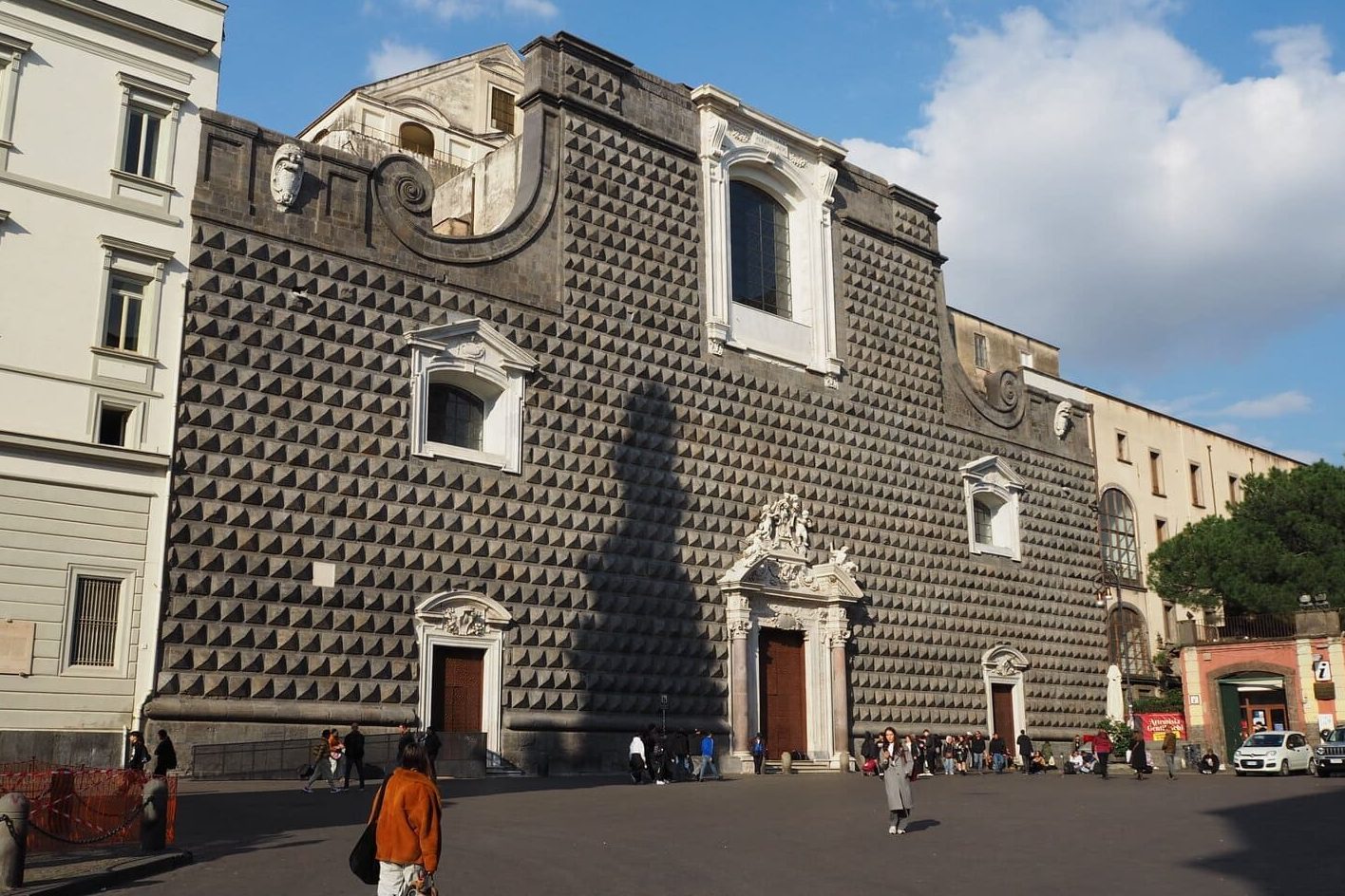 People walk in front of a large, dark stone building with a patterned facade and arched windows under a blue sky, one of the best places in Naples to admire historic architecture.