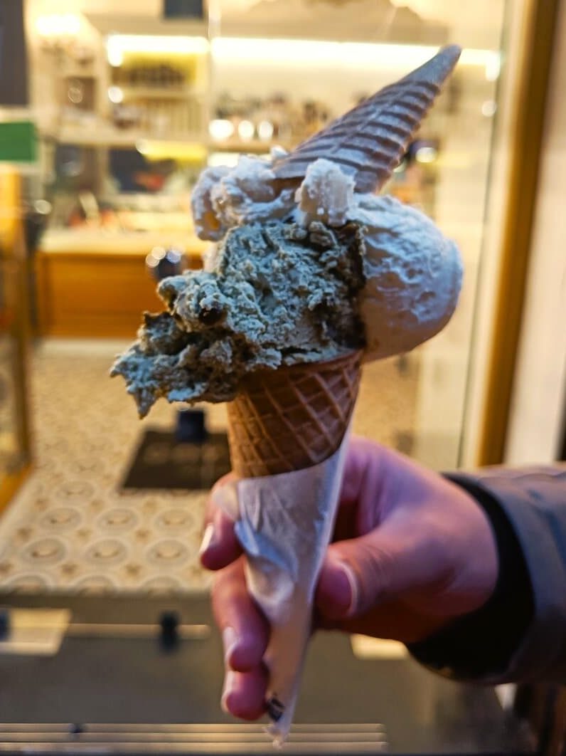 A hand holding a double-scoop ice cream cone with two flavors, in a shop with warm lighting—a delicious treat inspired by must try Naples food.