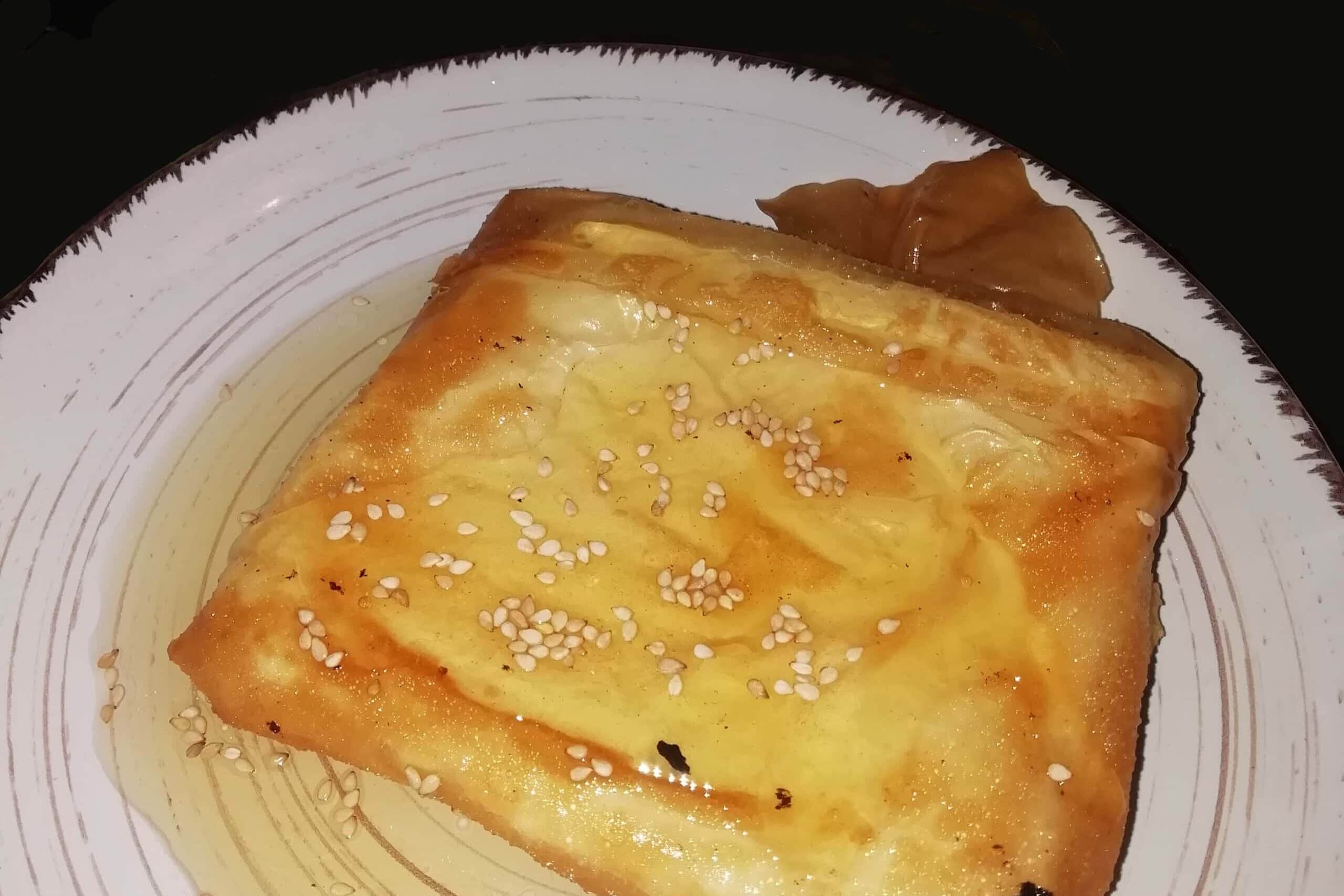 A square piece of honey-drizzled fried pastry topped with sesame seeds on a white plate—truly a must-try Greek food and one of the best comfort foods Athens has to offer.