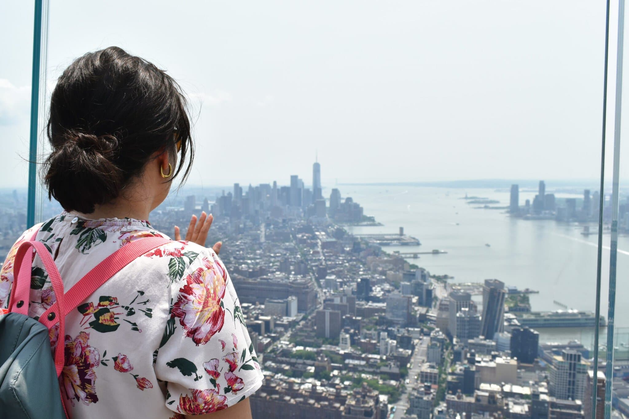 Woman with a floral rucksack looks out over a city skyline and river from a high vantage point.